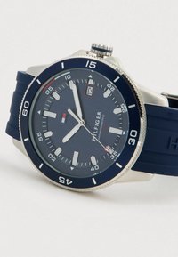 Tommy Hilfiger randkellaga sinise dials, hõbedaste markeritega, kuupäeva näidik, sinine raamis ja sinine silikoonist rihmaga valgel taustal.