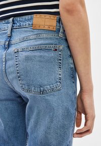 Kék farmernadrág barna Tommy Jeans címkével és kis Tommy Hilfiger zászlóval a hátsó zsebén, csíkos inggel viselve.