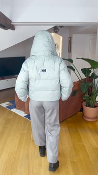 Puffig mintgrön jacka med stor huva, med en etikett på baksidan, kombinerad med grå byxor och svarta stövlar på ett trägolv.