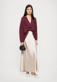 Bluză plisată burgundă cu decolteu în V și nasturi, asortată cu o fustă maxi fluidă din satin crem. Accesoriile includ o mică geantă de mână neagră.