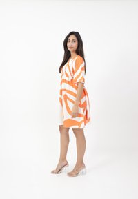 Vestido de patrón naranja y blanco con mangas cortas, escote en V y cintura ceñida, combinado con sandalias transparentes de tacón bloque.