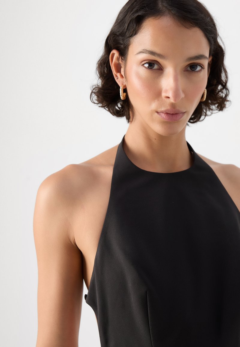 Czarna sukienka na halter z gładką fakturą i dopasowanym krojem, z odkrytymi bokami i minimalnymi szwami. Złote kolczyki w kształcie kół jako akcenty.