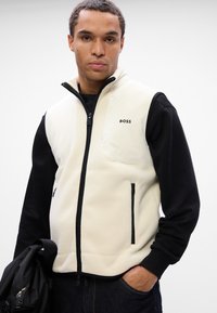 Γούνινο γιλέκο σε κρέμα με μαύρα μανίκια, διαθέτοντας ψηλό γιακά, κρυφές τσέπες με φερμουάρ και διακριτό λογότυπο της μάρκας στο στήθος. Μαλακή υφή και σύγχρονη γραμμή.