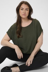 Oliivinvihreä, löysä yläosa, jossa on käännetyt hihat, yhdistettynä mustiin skinny-farkkuihin ja kiiltäviin mustiin kenkiin. Yksinkertainen, sileä pinta koko vaatteessa.