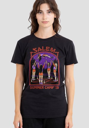 Young woman wears black T-shirt with illustration of five cheerleaders in shorts levitating a person above a pentagram, text "Salem Summer Camp '78."