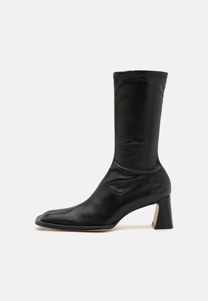 Fekete bőr bokacsizma, illeszkedő szárakkal, hegyes orral és vastag sarokkal. Simaságával és minimális varrási részleteivel rendelkezik.