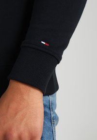 Polsino di maglione nero con trama a coste e piccolo logo con colori rosso, bianco e blu. Mano visibile e jeans in denim sullo sfondo.