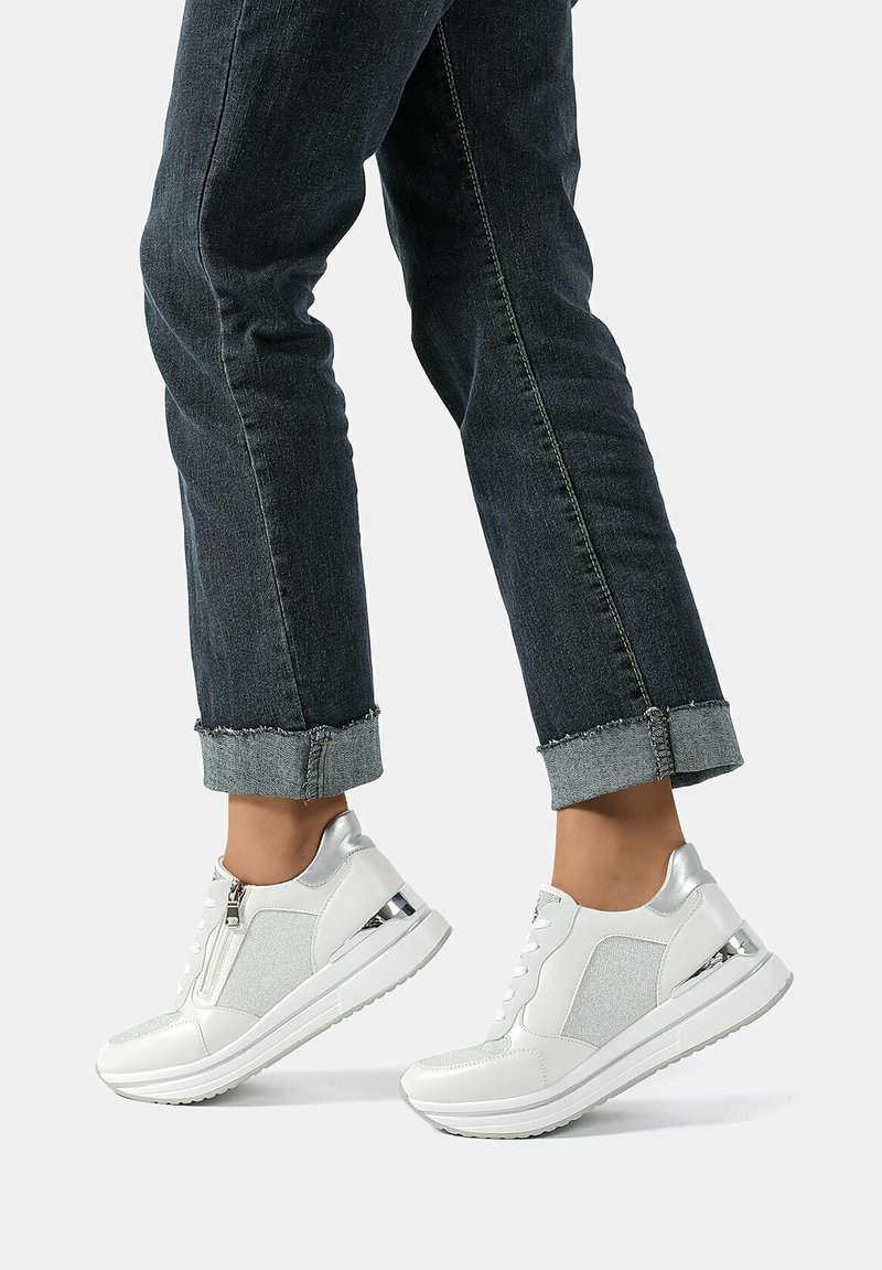 Sneakers bianchi con accenti metallici, chiusura a zip e pannelli laterali testurizzati. Abbinati a jeans scuri arrotolati per un look casual.