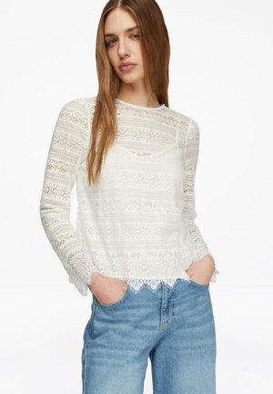 Młoda kobieta w białej koronkowej bluzce z długim rękawem i jasnoniebieskich dżinsach, z rękami w kieszeniach, stojąca na białym tle.