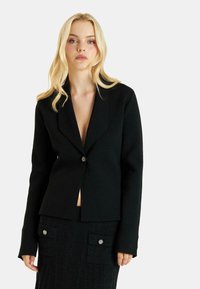 Blazer negro con solapas con muesca, cierre de un solo botón y mangas largas; combinado con una falda acanalada que presenta bolsillos frontales y acentos de botones.