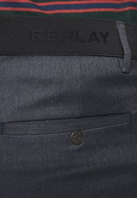 Pantalons en denim noir avec une texture tissée, ceinture élastique noire avec logo "REPLAY" en relief et fermeture à bouton sur la poche arrière.