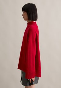 Roter, übergroßer Pullover mit hohem Rippkragen, weiten Ärmeln und geripptem Saum. Kombiniert mit einem schwarzen Lederrock.