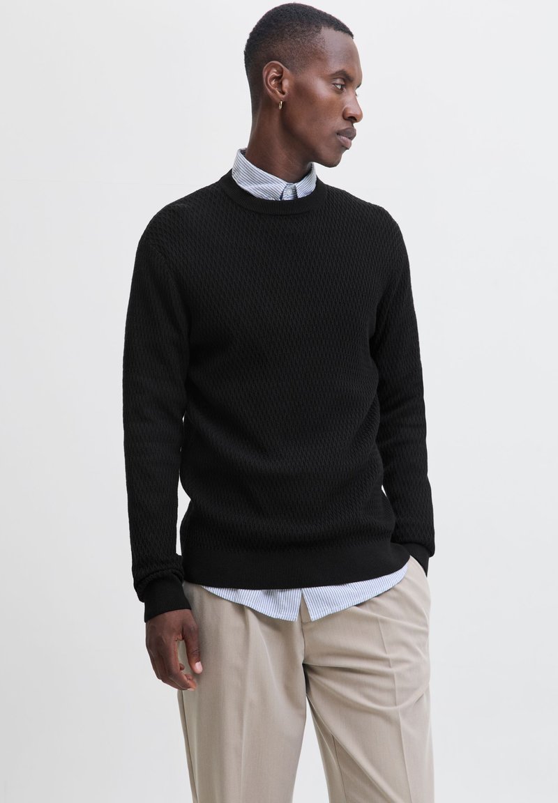 Jeune homme portant un pull noir par-dessus une chemise rayée bleue avec un pantalon beige, regardant sur le côté devant un fond clair uni.