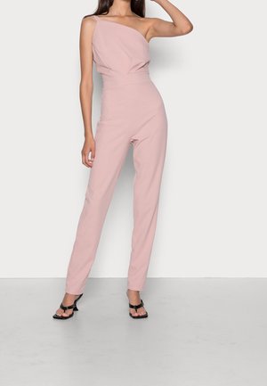 Žena v růžovém jednokrajném overal s jedním raménkem a černých sandálech na podpatku, stojící s jednou rukou zvednutou ke hlavě.