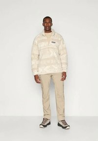 Fleece genser i kremfarge med geometriske mønstre, halv-zip krage, merketapet, kombinert med lysebeige bukser og turtøfler.