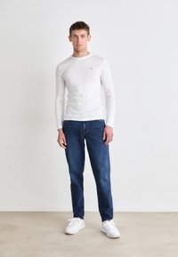 Wit langemouwen shirt, blauwe denim jeans en witte sneakers. Het shirt is slim fit, met een ronde halslijn en subtiele logo-details.