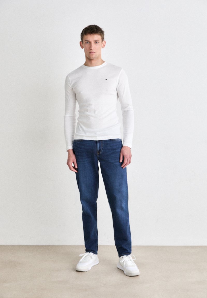 Wit langemouwen shirt, blauwe denim jeans en witte sneakers. Het shirt is slim fit, met een ronde halslijn en subtiele logo-details.