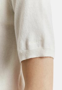 Gestickter weißer kurzärmeliger Pullover mit glatter Textur, gerippten Bündchen und einem dezenten Logo-Detail nahe dem Ärmelabschluss.