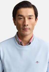 Homme aux cheveux noirs courts portant un sweat-shirt bleu clair par-dessus une chemise rayée à col, face à l'avant avec une expression neutre sur fond blanc.