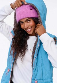 Modrý zateplený vest s šedou fleecovou podšívkou, s růžovými zipovými detaily. Model má na sobě růžovou pletenou čepici a bílou mikinu.