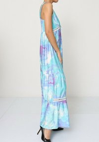 Robe maxi en motif tie-dye bleu et violet, avec un corsage froncé, des dentelles et une jupe à volants. Portée avec des escarpins noirs.
