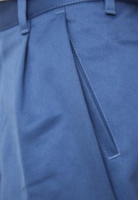 Pantalones de tela azul con frente plano y un sutil detalle de pliegue. El material es suave, con costuras precisas y un acabado limpio.