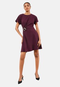 Beerenlila Kleid mit kurzen Ärmeln, gedrehtem Vorderteil und silberfarbener Gürtelschnalle. Glatter Stoff, knielang mit schwingendem Rock.