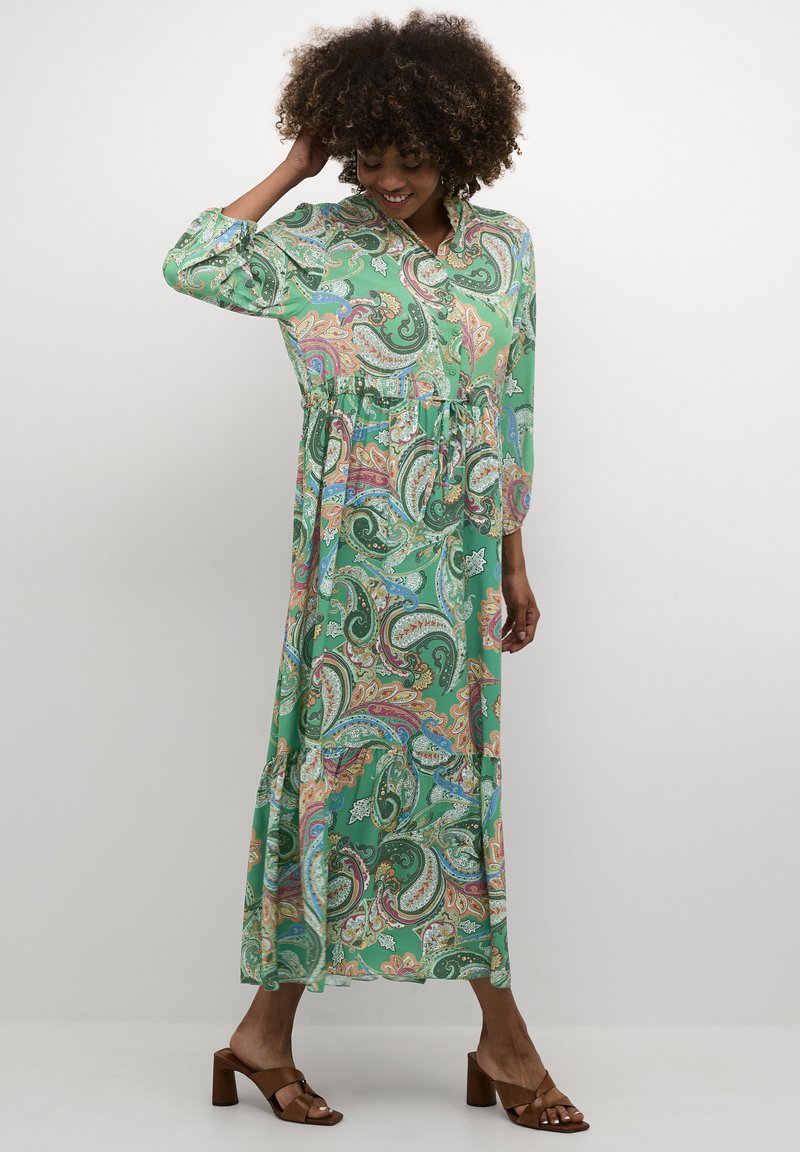Zeleni maksi oblek z vzorcem paisley, z ovratnikom na gumbke, nabranim pasom in dolgimi rokavi. Model nosi rjave sandale s peto.