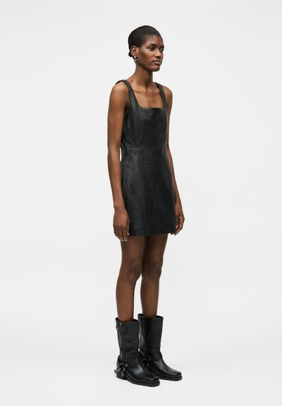 Γυναίκα που στέκεται φορώντας ένα μίνι μαύρο δερμάτινο φόρεμα χωρίς μανίκια σε συνδυασμό με μαύρα δερμάτινα μποτάκια μέχρι τη μέση της μοσχαρίδας με λεπτομέρειες από πόρπες και αλυσίδες.