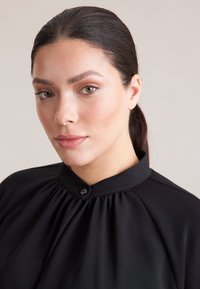 Blusa nera con un design plissettato e chiusura con bottone al collo. Tessuto liscio con una leggera lucentezza. Primo piano del collo e delle spalle.