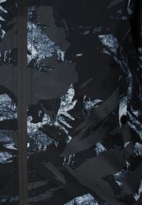 Zwarte stoffen jas met een getextureerd abstract patroon in grijs en wit. Voorzien van een ritssluiting en een drukknoopdetail.