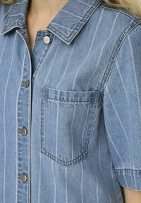 Helles blaues Jeanshemd mit vertikalen weißen Streifen, kurzen Ärmeln, Kragen und einer Brusttasche. Verfügt über metallische Druckknöpfe.
