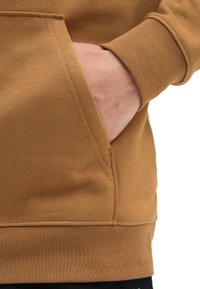 Sudadera de algodón marrón con un bolsillo tipo canguro y puños acanalados. Tejido texturizado con detalles de costura visibles. Cintura negra visible por debajo.