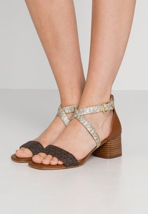Pieds portant des sandales à talons marron avec des brides croisées beige clair et une boucle dorée sur un fond neutre.
