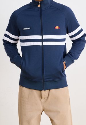 Man draagt een marineblauwe jas met rits met witte horizontale strepen en beige broek, handen in de jaszakken.