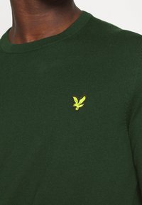 Gros plan d'une personne portant un pull en maille vert foncé avec un petit emblème d'oiseau jaune sur le côté gauche de la poitrine.
