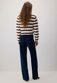 Persoană cu păr lung și creț, purtând un pulover în dungi maro și alb și blugi largi de culoare albastră închisă, stând în fața unui perete simplu de o culoare deschisă.