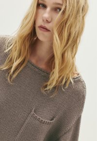 Junge Frau mit welligem, blonden Haar, die einen locker gestrickten, taupefarbenen Pullover mit einer kleinen Brusttasche trägt und leicht von der Kamera wegschaut.
