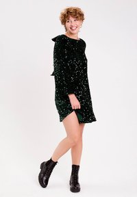 Jeune personne avec des cheveux courts et bouclés, portant une robe en sequins vert foncé et des bottines noires à lacets, souriant et regardant sur le côté.