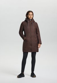 Brauner Puffermantel mit Kapuze, Reißverschluss vorne und gerippter Textur. Er hat eine schlanke Passform und wird mit schwarzen Leggings und Schuhen kombiniert.