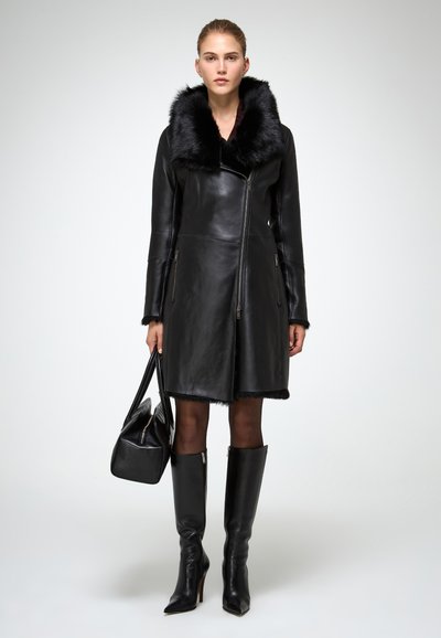 Donna con un mantello di pelle nera con colletto di pelliccia, collant neri trasparenti, stivali neri al ginocchio e con una borsa nera su uno sfondo bianco.