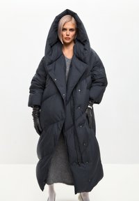 Padded crni kaput s velikom kapom, oversize kroj i zakopčavanje na dugmad. Karakterizira ga pokrivač s kvadratićima i bočne džepove.