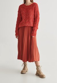 Czerwony sweter z teksturowanym splotem, z opadającymi ramionami, zestawiony z plisowaną midi spódnicą w kolorze rdzy oraz jasnobeżowymi botkami na sznurówki. 