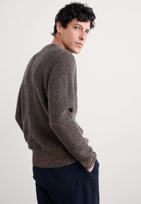 Brązowy sweter dzianinowy o prążkowanej fakturze, z okrągłym dekoltem i długimi rękawami, zestawiony z granatowymi spodniami, pokazany z boku z lekkim obrotem.