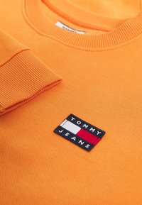 Oranžová bavlněná mikina se žebrovaným výstřihem a manžetami. Obsahuje černou nášivku s nápisem "TOMMY JEANS" a červenobílým vzorem.