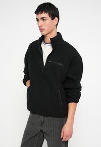 Νέος άνδρας που φοράει μαύρο φλις πουλόβερ με φερμουάρ στο στήθος και πλαϊνές τσέπες, στέκεται με τα χέρια στις τσέπες, κοιτάζοντας προς τα αριστερά σε λευκό φόντο.