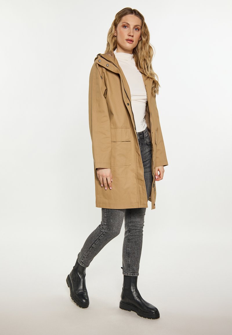 Femme portant un manteau à capuche beige, un col roulé blanc, un jean gris et des bottines noires, se tenant sur un fond blanc uni.