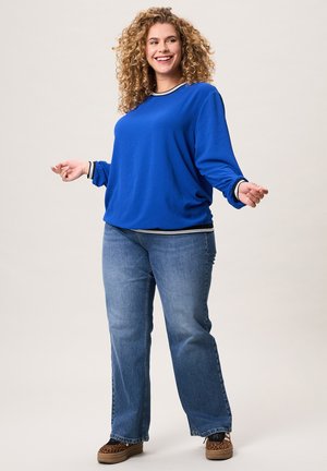 Vrouw met krullend haar die glimlacht, gekleed in een blauw shirt met lange mouwen, blauwe jeans en schoenen met luipaardprint, staand tegen een effen achtergrond.