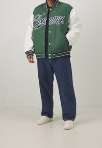 Zielono-biała kurtka varsity z logo "Tommy", ściągaczem na kołnierzu, guzikami i paskami dekoracyjnymi. Noszona na ciemnych dżinsach i białych trampkach.