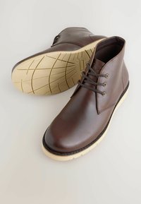 Pair of dark brown leather ankle boots with beige rubber soles and brown laces, one boot showing tread pattern underneath.